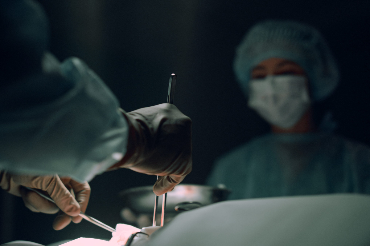 Gloved hands of a surgeon holding instruments over a patient in a dim operating room.