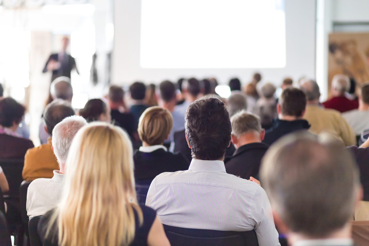 A group of sitting people watching a person blurred out in the forground giving a presentation.